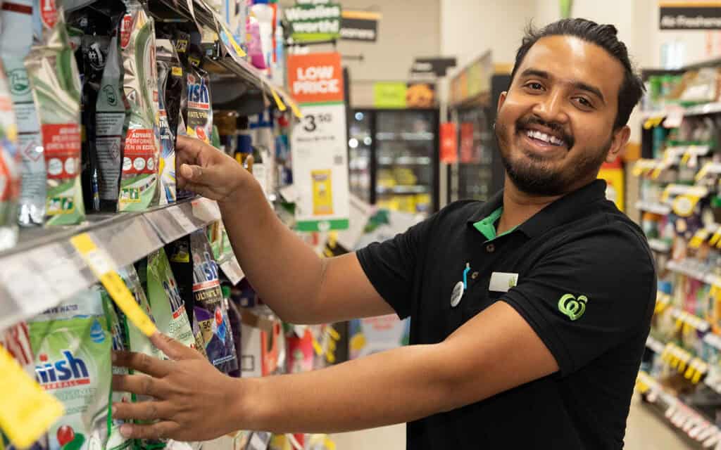 A Woolworths worker is smiling while stacks the shelves in the supermarket aisle.